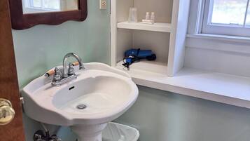 Et badeværelse med et pedalvask, et spejl, et vindue med skodder og en hylde med toiletartikler.