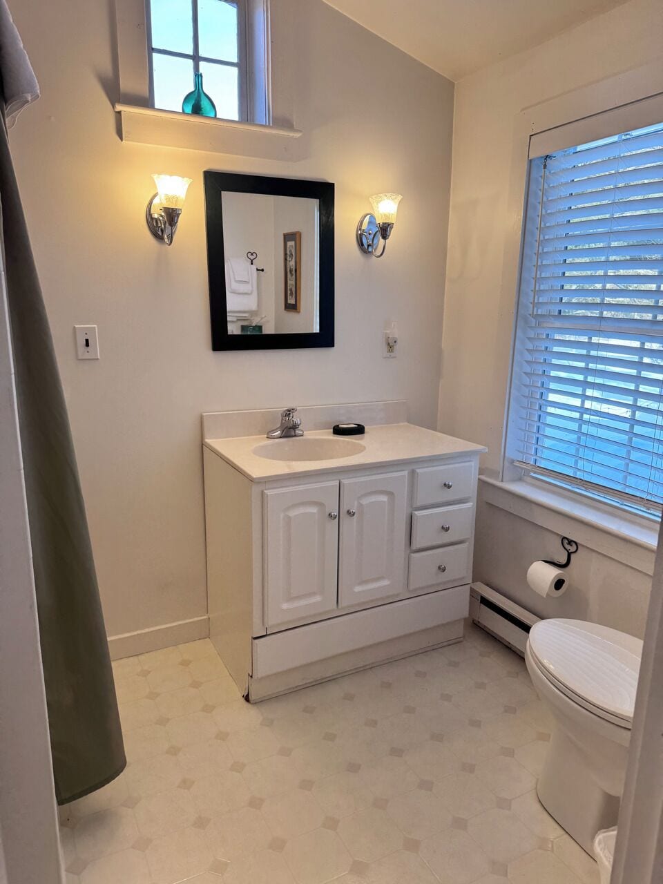 A bathroom with a sink, mirror, two wall sconces, a toilet, and a window with blinds.