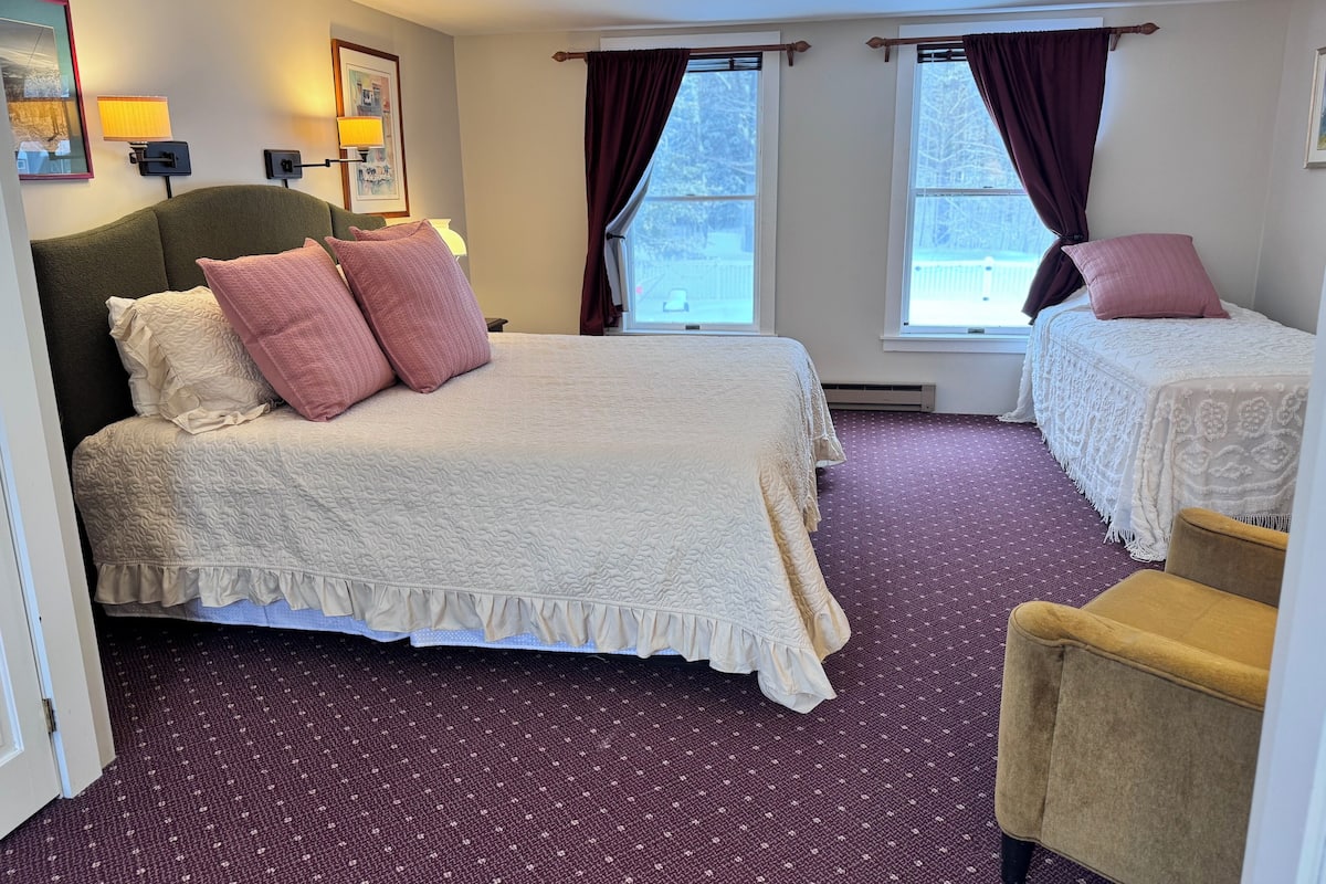 A hotel room with a large bed, two smaller beds, a chair, and a window with curtains.