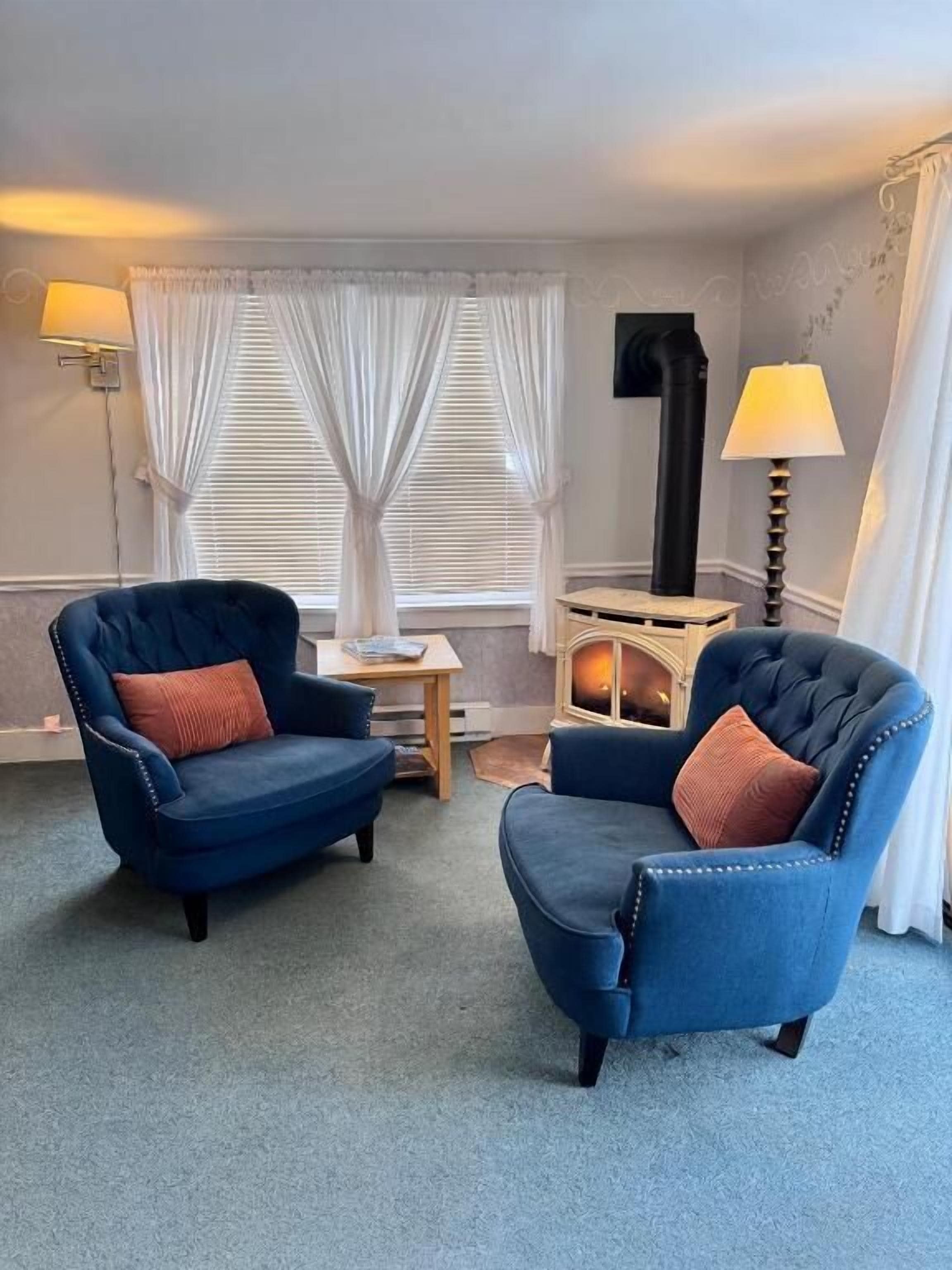 A room with two blue armchairs, a small table, a fireplace, and a lamp.