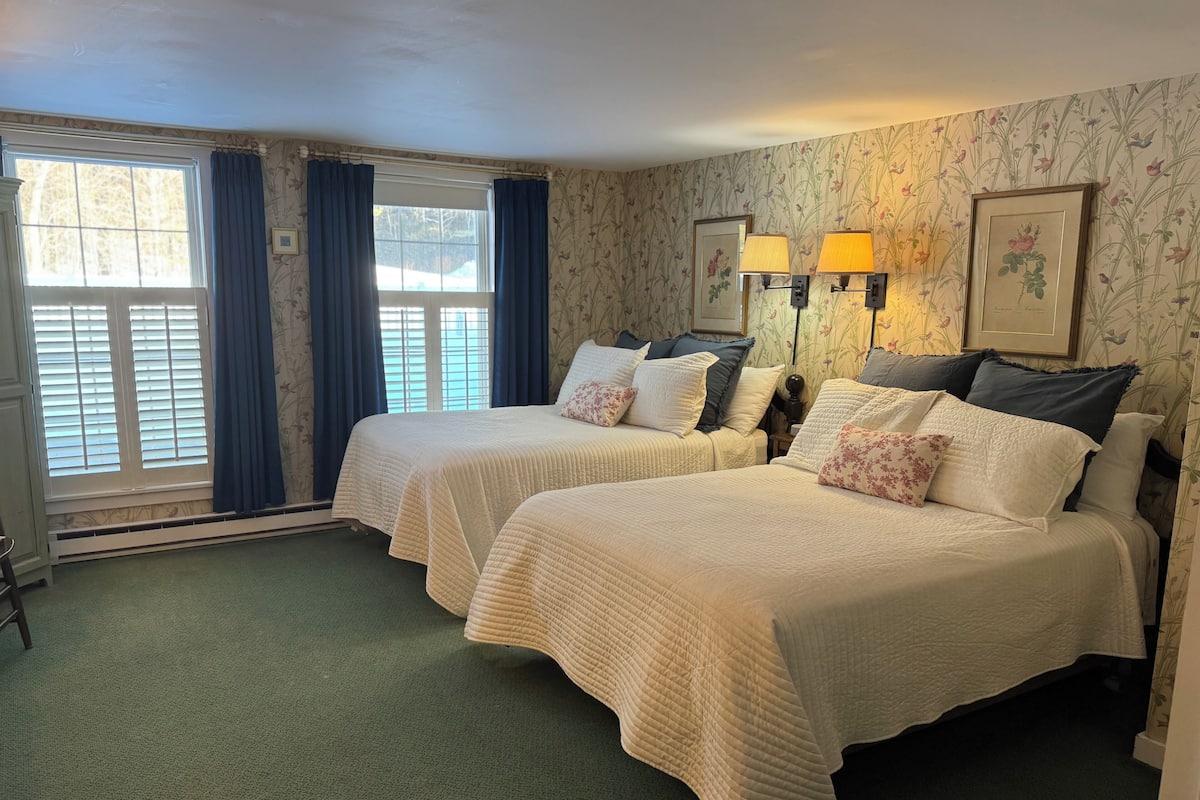 A hotel room with two beds, a window with curtains, a radiator, and a wall with floral wallpaper.
