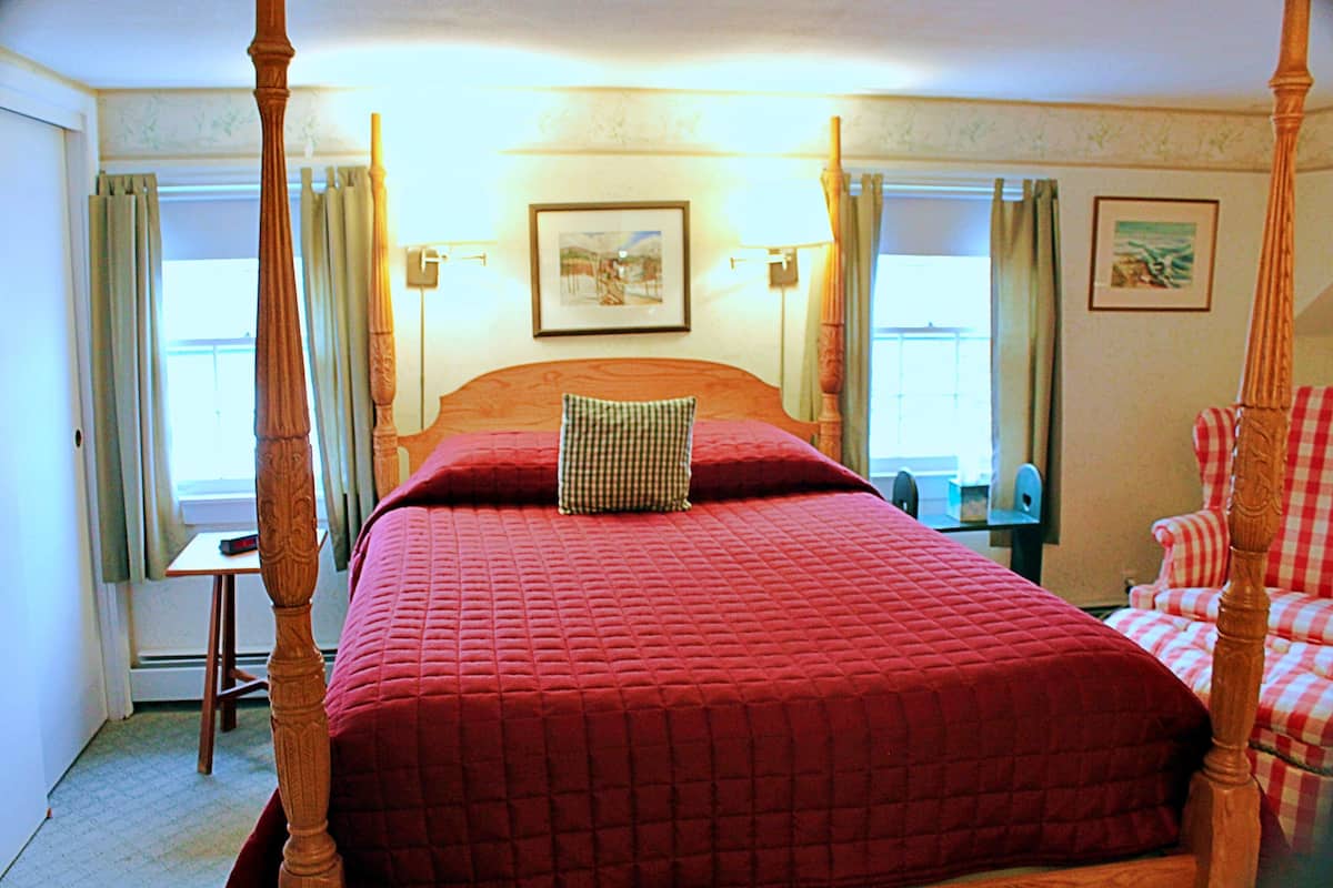A four-poster bed with a red quilt, a checkered pillow, and a wooden headboard. There is a small table and a chair to the left, and a checkered chair to the right.