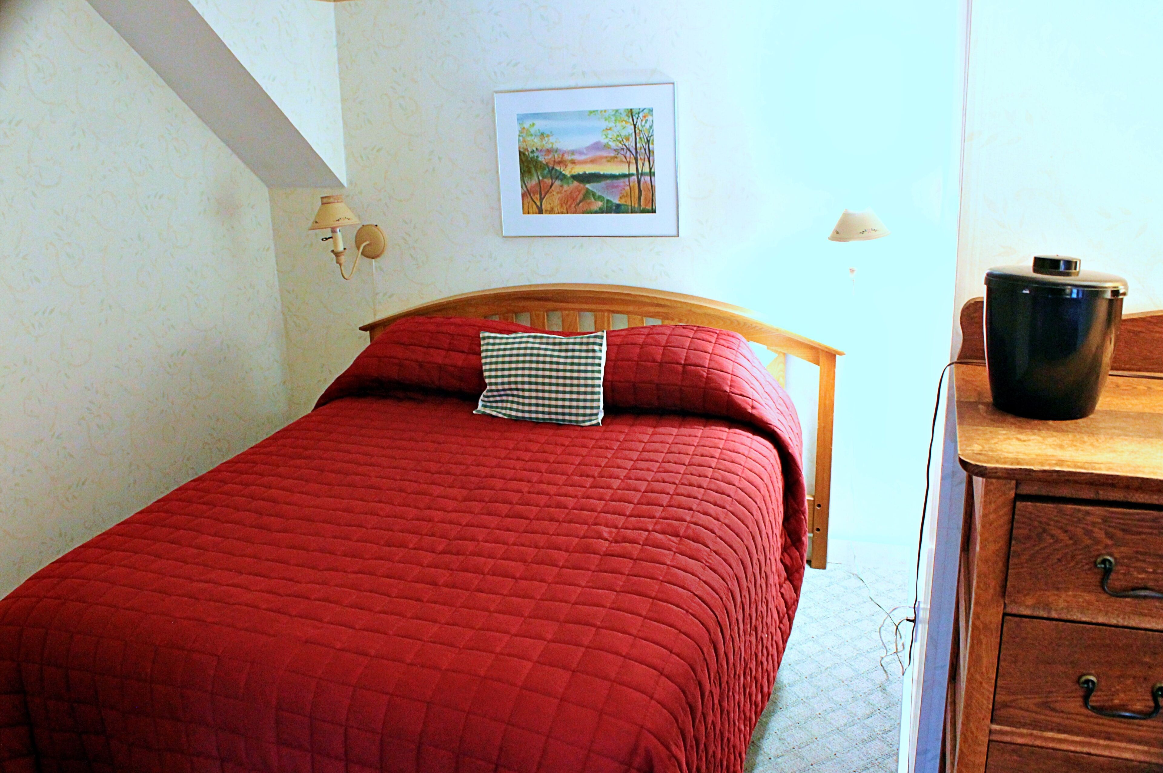 A bed with a red quilt, a wooden headboard, a checkered pillow, a framed landscape painting on the wall, and a wooden dresser with drawers.