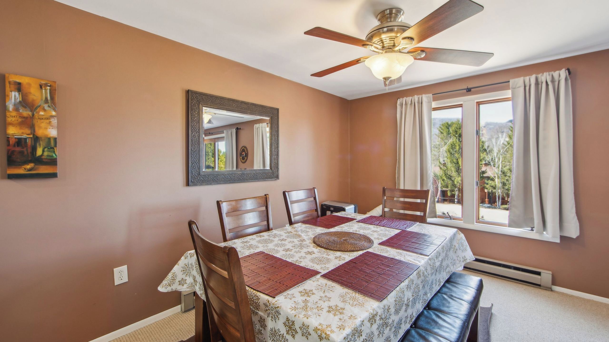 A dining room with a table, chairs, a ceiling fan, a window with curtains, and a painting on the wall.