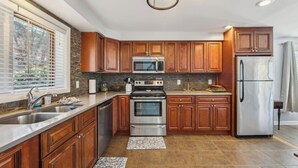 A modern kitchen with wooden cabinets, stainless steel appliances, and a large window with blinds. - Hullabaloo by Avantstay Indoor Heated Pool + Game Room Near Gore Mountain (North Creek)