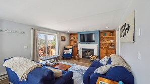 A living room with a fireplace, a television, and a view of the outdoors. - Hullabaloo by Avantstay Indoor Heated Pool + Game Room Near Gore Mountain (North Creek)