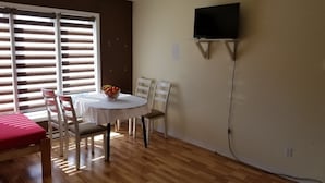 Uma sala de jantar com uma mesa redonda, cadeiras brancas, um sofá vermelho e uma TV fixada na parede.