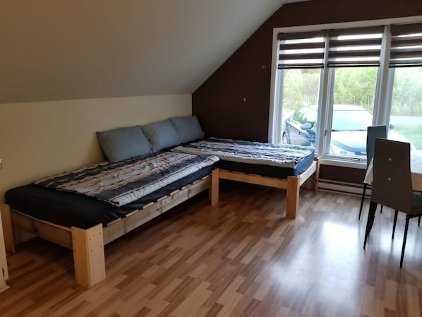 A loft-style room with a wooden floor, a bed with a patterned bedspread, a window with blinds, and a chair.