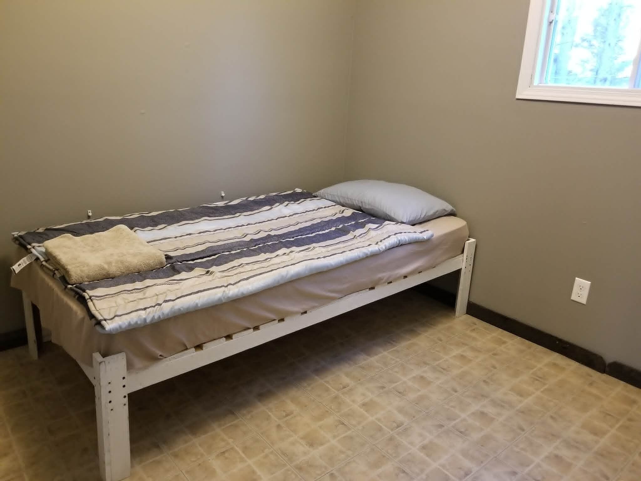 Una cama individual con una colcha y almohada a rayas, una mesita de noche pequeña y una ventana con vistas a árboles.