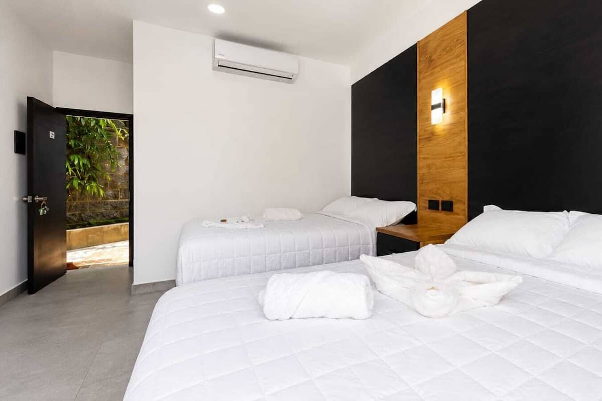 Ett hotellrum med två sängar, en vägg med träpanel som ger ett snyggt intryck, en luftkonditioneringsanläggning och utsikt över grönområden genom en öppen dörr.