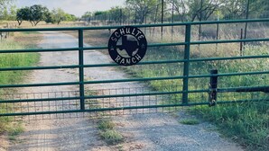A gate with a logo reading 'Schulze Ranch' and a gravel path leading through a fenced area with trees and grass.