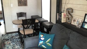 A living room with a gray sofa, teal pillows, a patterned rug, a dining area with a table and chairs, and a mounted TV.