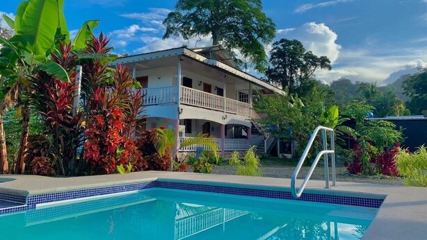 A pool with a tiled edge, a two-story building with balconies, and tropical vegetation.