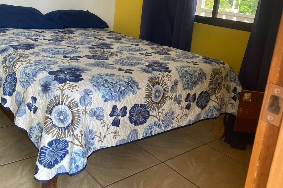 Uma cama com colcha florida, uma janela com cortinas e piso ladrilhado.