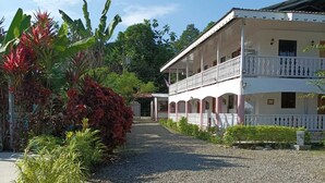 A two-story building with a balcony, surrounded by tropical plants and a gravel pathway.