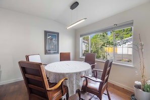 A dining area with a round table, wooden chairs, a framed picture on the wall, and a window with a view of greenery.