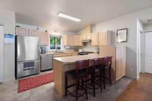 A modern kitchen with a stainless steel refrigerator, wooden cabinets, a central island with bar stools, and a red rug.