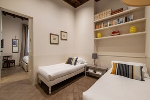 Ein Hotelzimmer mit zwei Betten, einem kleinen Tisch, einem Regal mit Büchern und Dekoration sowie einer Tür zu einem weiteren Raum.