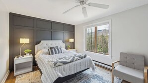 Un dormitorio con cama, dos mesitas de noche, una mesita auxiliar, una silla, un ventilador de techo y una ventana con vistas.