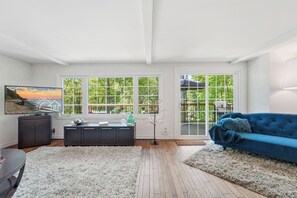 Ein Wohnzimmer mit einem blauen Sofa, einem Fernseher, einem Schreibtisch und großen Fenstern mit Blick auf Grünflächen.