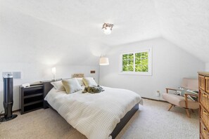 Ein Schlafzimmer mit Bett, Nachttisch, Lampe und einem Fenster mit Blick auf Grünflächen.