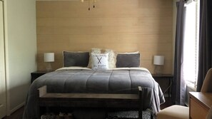 Ein Bett mit Kopfteil, zwei Nachttische mit Lampen, ein Kopfkissen mit Logo und ein Stuhl zur rechten Seite.