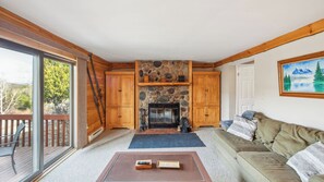 A cozy living room with a stone fireplace, wooden cabinets, a sofa, and a balcony with a view of trees. - Echo by Avantstay Sauna + Indoor Pool + Fireplace Near Gore Mountain (North Creek)