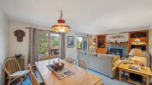 A cozy living room with a fireplace, a dining area with a wooden table, and a large window offering a view of the outdoors. - Quick Silver by Avantstay Sauna + Heated Pool + Game Room Near Gore Mtn (North Creek)