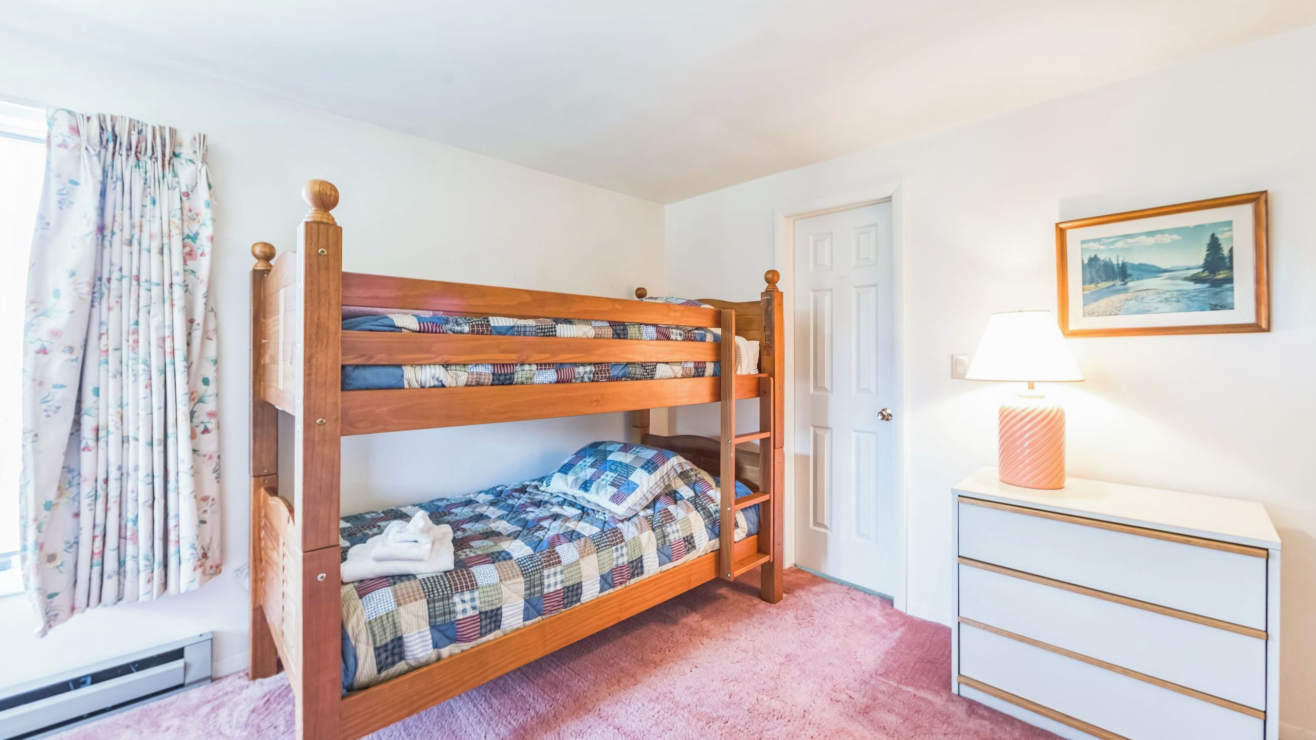 Una cama litera con colcha a cuadros, una mesita de noche con lámpara, un cuadro enmarcado en la pared y una ventana con cortinas florales.