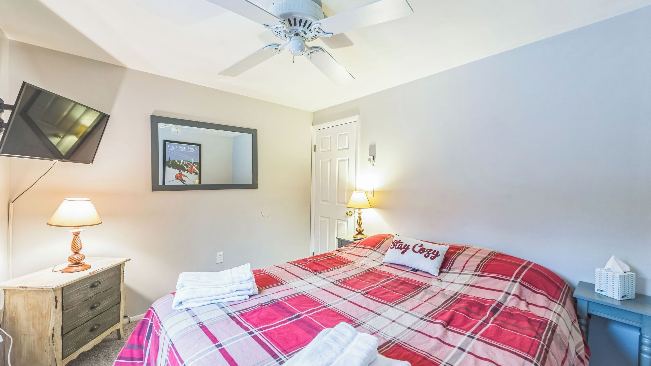 Ein Schlafzimmer mit einem Bett, einem Deckenventilator, einem Fernseher, einem Spiegel und einem Nachttisch mit Lampe.