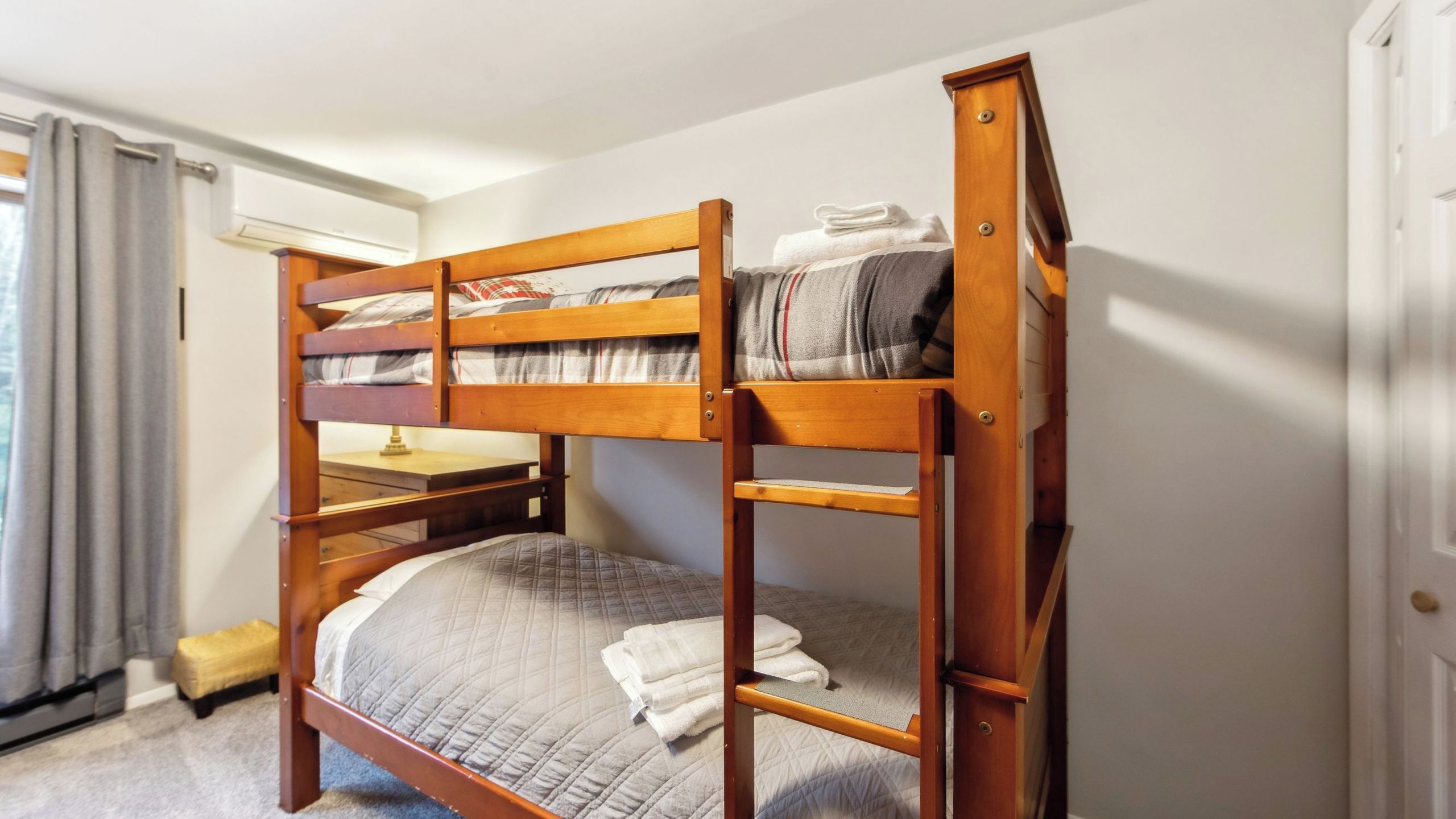 A bunk bed with a desk and chair, a small stool, and a window with curtains.