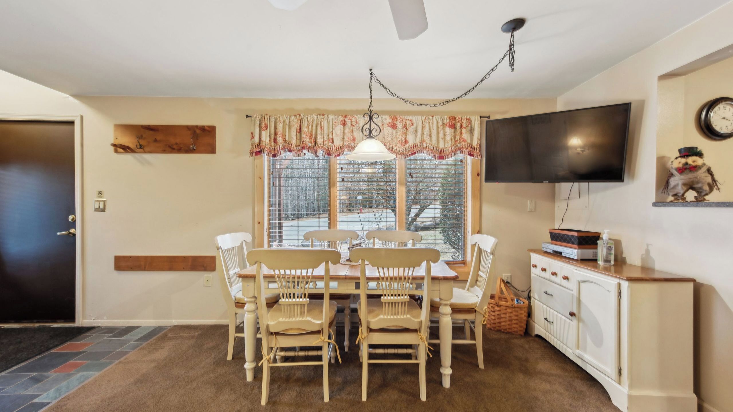 A dining area with a table and chairs, a television on the wall, and a window with blinds.
