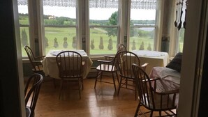 A dining area with a table and chairs, large windows with lace curtains, and a view of a green landscape. - New Glasgow Inn (New Glasgow)
