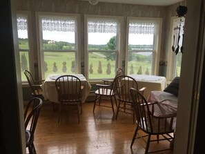Ein Essbereich mit Tisch und Stühlen, große Fenster mit Spitzendeckchen und Blick auf eine grüne Landschaft.