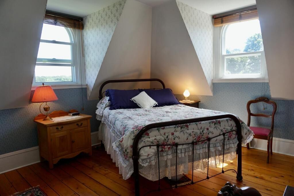 A bedroom with a bed, nightstand, lamp, chair, and two windows.