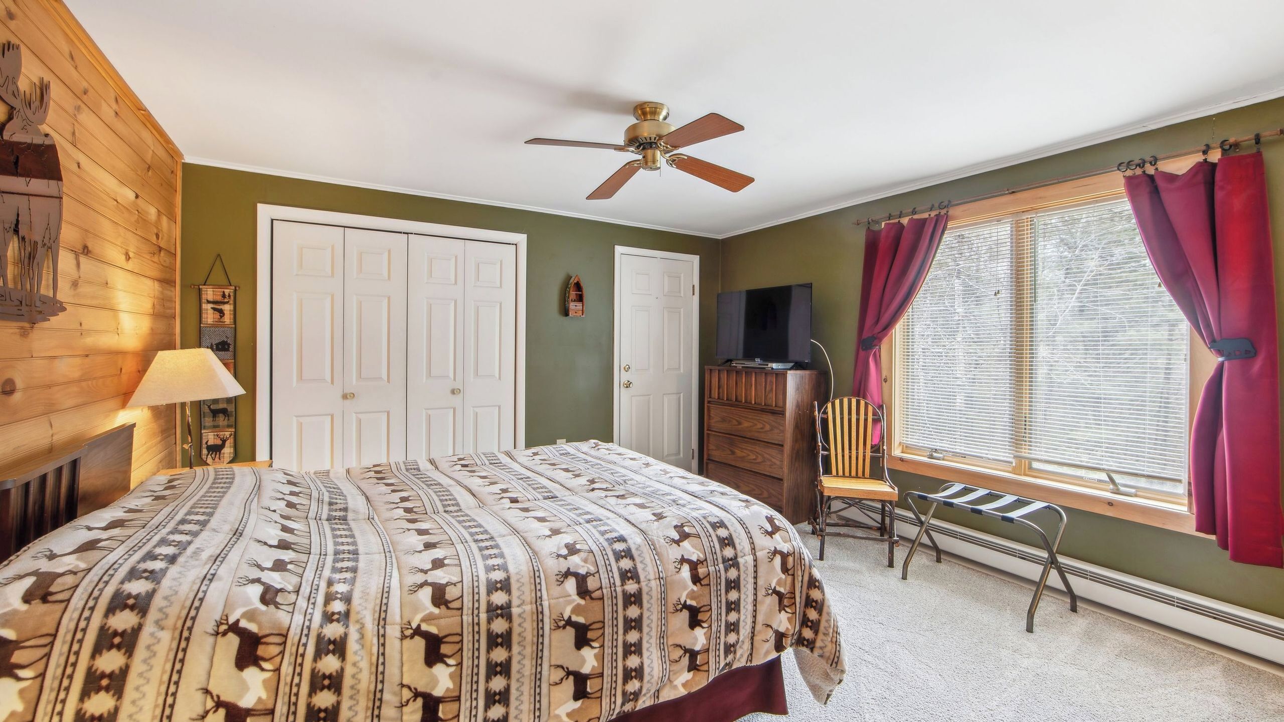 A bedroom with a bed, a ceiling fan, a dresser, a chair, a window with curtains, and a wall-mounted lamp.