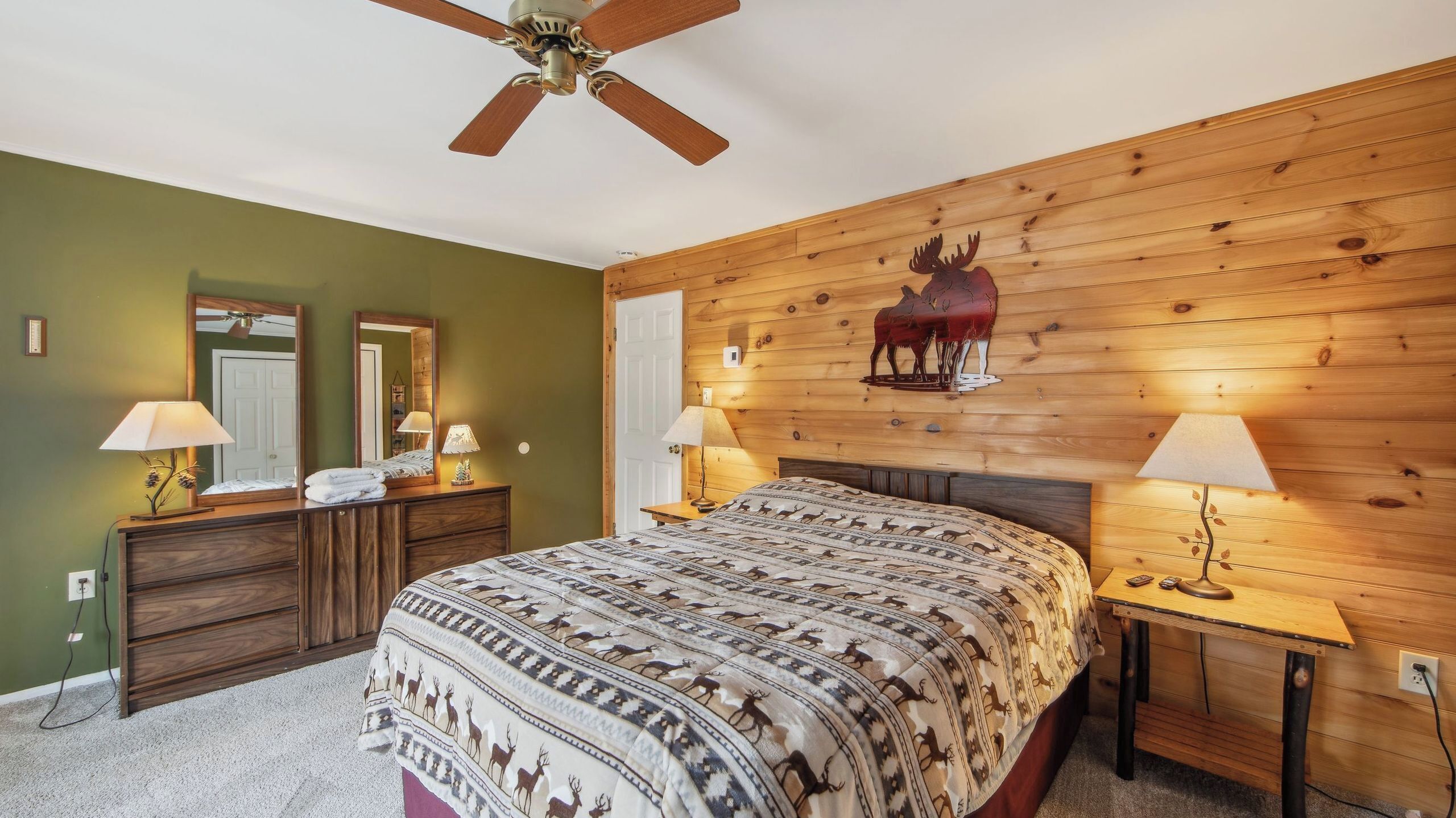 Een slaapkamer met een houten hoofdeinde, een bed met een geborduurd plaid, twee bedlampen, een plafondventilator en een houten muur met een afbeelding van een eland.