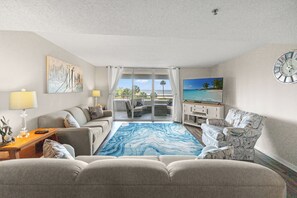 Ein Hotelzimmer mit einer Couch, Sesseln, einem Fernseher, einer Uhr und Blick auf den Ozean durch eine Schiebetür.