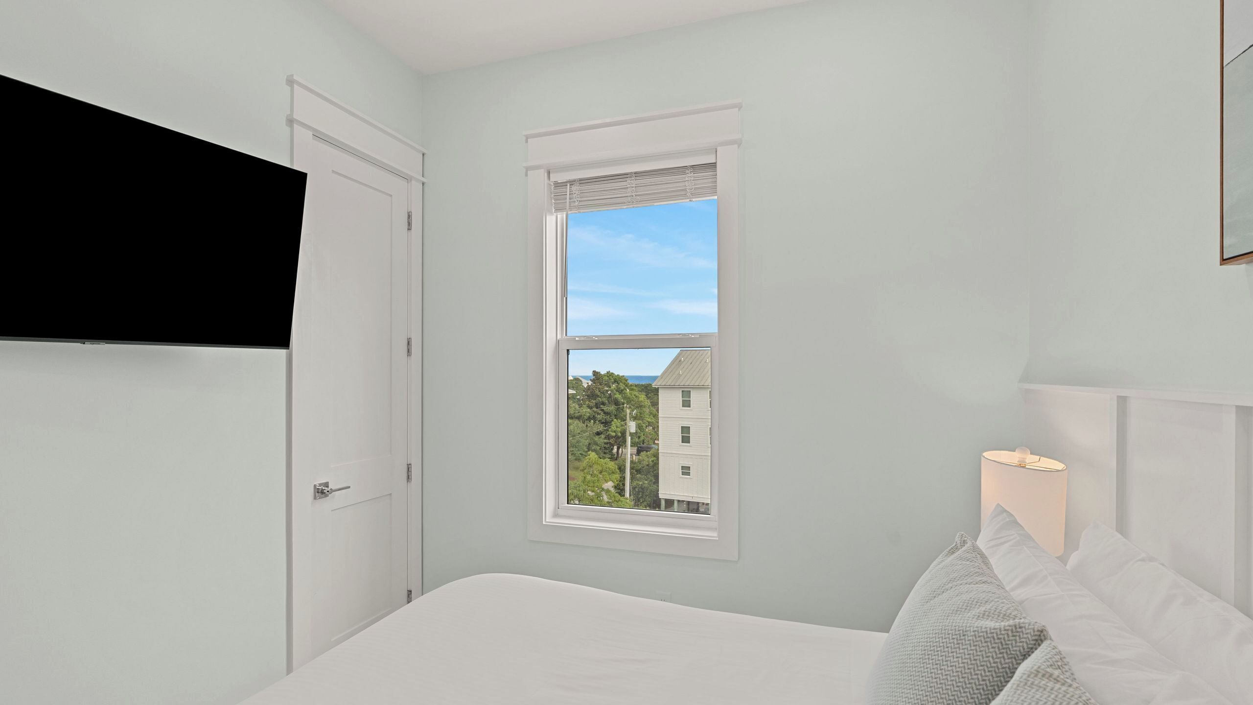 Une chambre à coucher avec un lit, une fenêtre donnant sur des immeubles, une porte et un téléviseur fixé au mur.