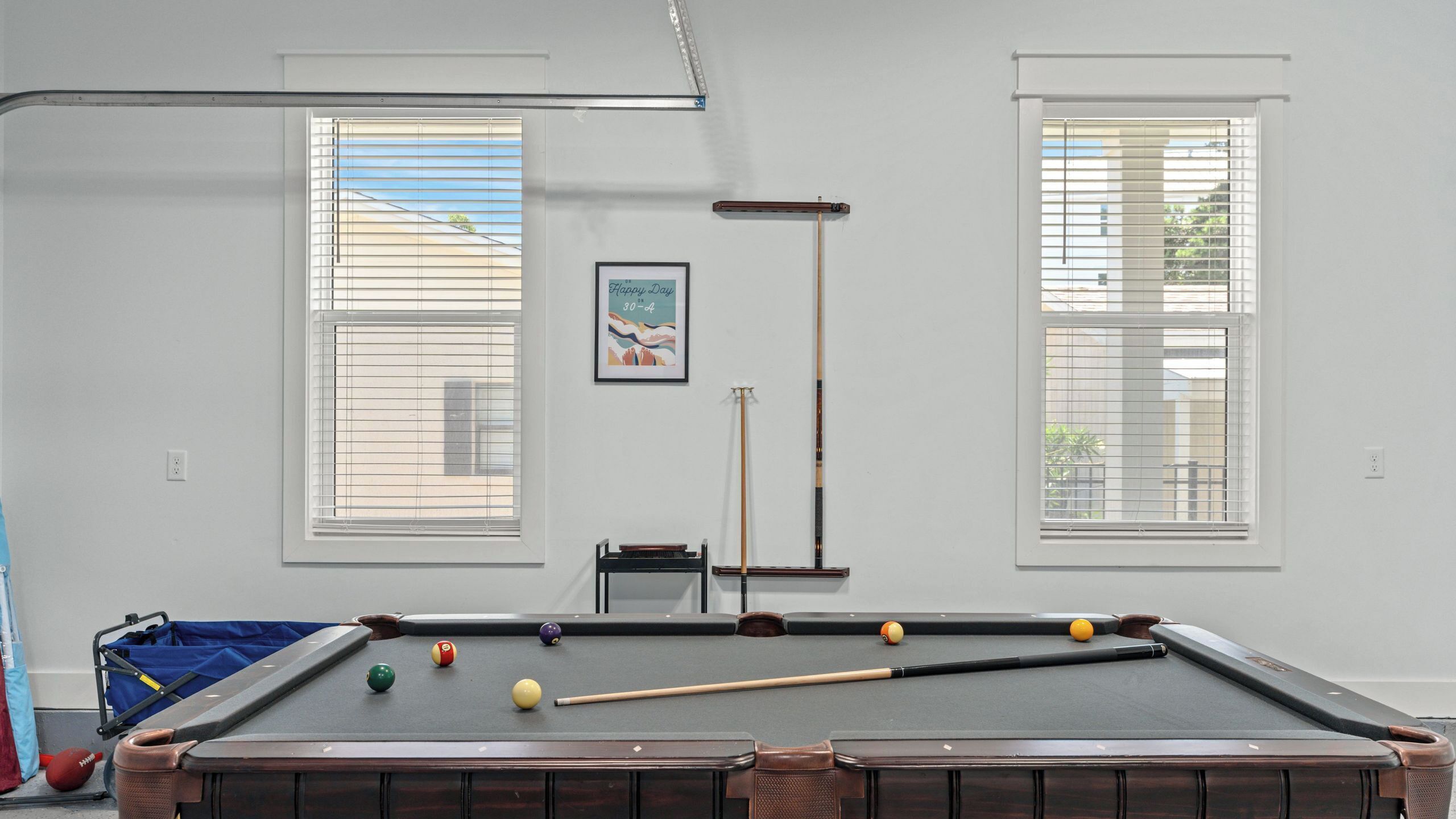 Ein Billardtisch mit Kugeln und Spielstöcken, Fenster mit Jalousien und ein gerahmtes Bild an der Wand.