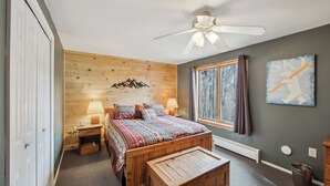 A bedroom with a wooden headboard, a bed with patterned bedding, a nightstand with lamps, a wooden bench, a ceiling fan, a window with a view of trees, and a wall art piece. - Cloud by Avantstay Sauna + Indoor Heated Pool + Game Lounge Near Gore Mtn (North Creek)
