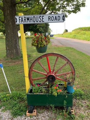 Un cartel de Farmhouse Road, una maceta con forma de rueda de madera con flores, y una jardinera verde.