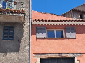 Ein Gebäude mit strukturierter Fassade, einem Balkon mit Topfpflanzen und einem Fenster mit Rollläden.