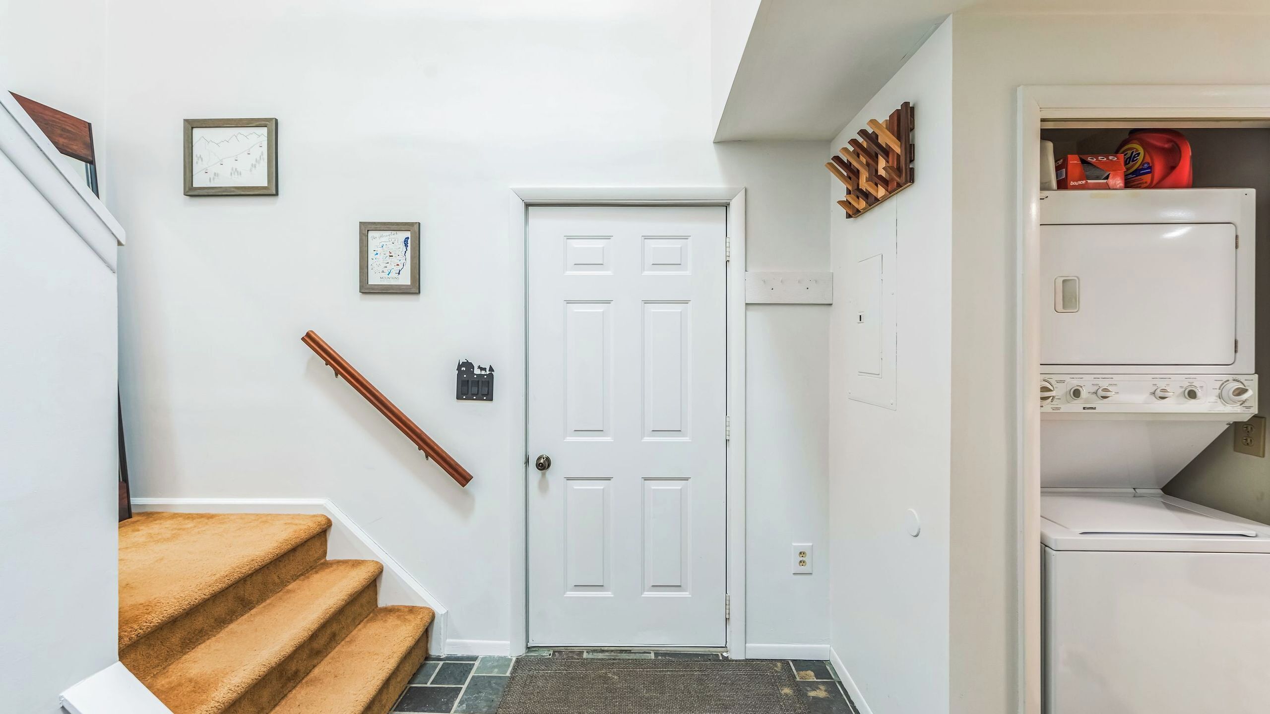 A hallway with a white door, a staircase with a brown carpet, and a laundry area with stacked items.
