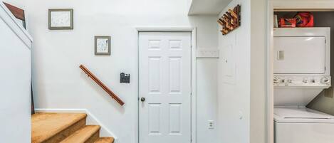 A hallway with a white door, a staircase with a brown carpet, and a laundry area with stacked items.