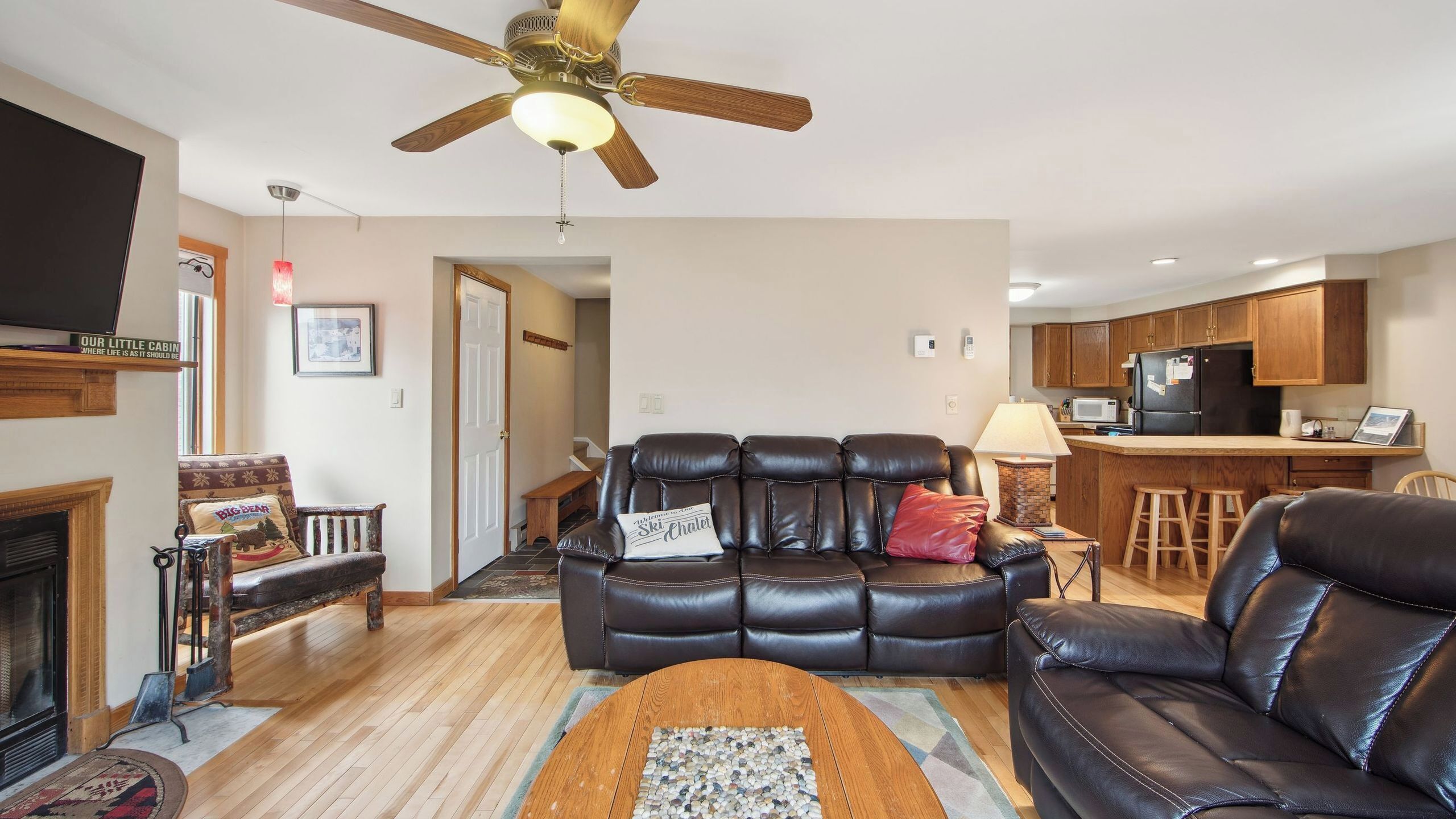 A living room with a leather sofa, a fireplace, a ceiling fan, and a kitchen area with wooden cabinets.