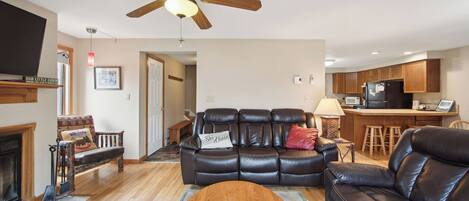 A living room with a leather sofa, a fireplace, a ceiling fan, and a kitchen area with wooden cabinets.