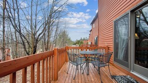 A wooden deck with a table and chairs, a glass door, and a view of trees and buildings. - Chatiemac by Avantstay Wood Fireplace + Resort Pool + Game Room Ski Retreat (North Creek)