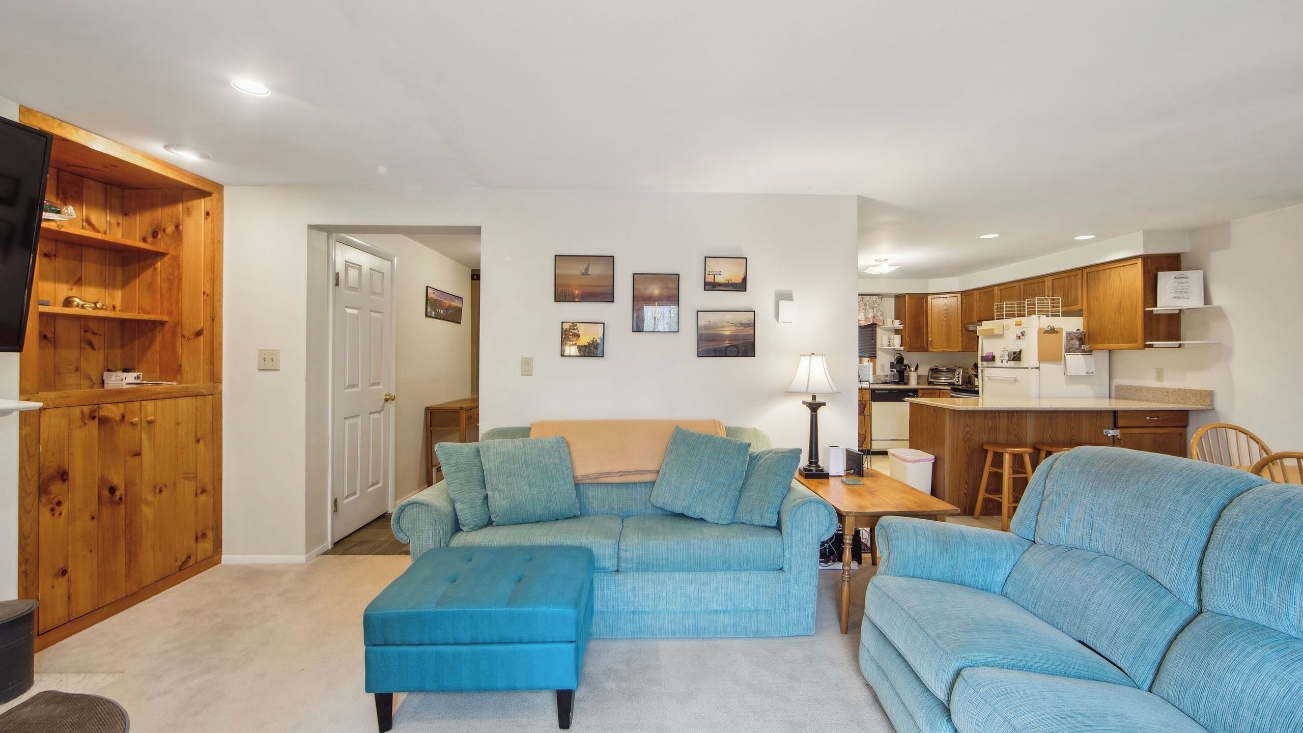 A living room with a sectional sofa, a wooden cabinet, and a kitchen area with wooden cabinets and a refrigerator.