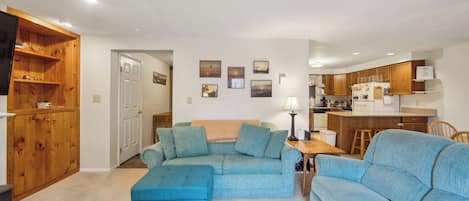 A living room with a sectional sofa, a wooden cabinet, and a kitchen area with wooden cabinets and a refrigerator.